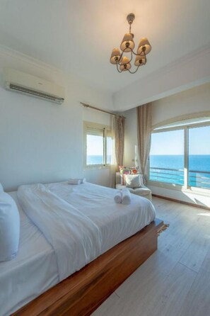 A bedroom with a large bed, a ceiling light fixture, a window with curtains, and a view of the sea.