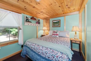 A bedroom with a wooden ceiling, a bed with floral bedding, two bedside lamps, a window with white curtains, and a shelf with decorative items. - NEW!! Pondview Cabin - Private Dock on West Pond (Parsonsfield)