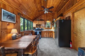A cabin interior with a wooden dining table, chairs, a kitchen with a stove, refrigerator, and a ceiling fan. - NEW!! Pondview Cabin - Private Dock on West Pond (Parsonsfield)