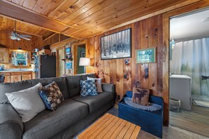 L’intérieur d’un chalet accueillant, avec un plafond en bois, un canapé gris agrémenté de coussins à motifs, un banc bleu et un coin repas comprenant un réfrigérateur et une lampe.