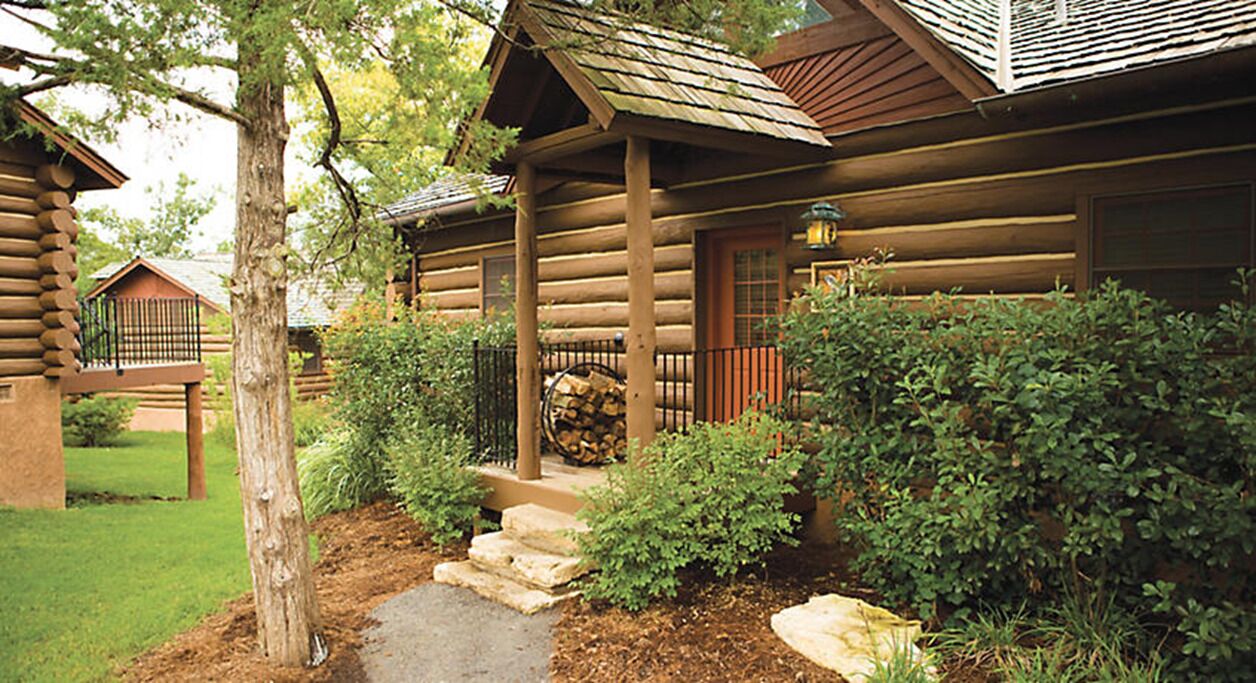 A log cabin with a stone pathway leading to the entrance, surrounded by greenery and a stack of firewood.