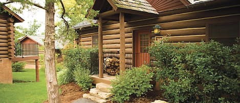 A log cabin with a stone pathway leading to the entrance, surrounded by greenery and a stack of firewood.