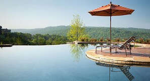 A clear swimming pool with a lounge chair and umbrella, surrounded by greenery and hills. - Celebrate Labor Day weekend in the Missouri Ozarks in this cabin for 6 guests. (Ridgedale)