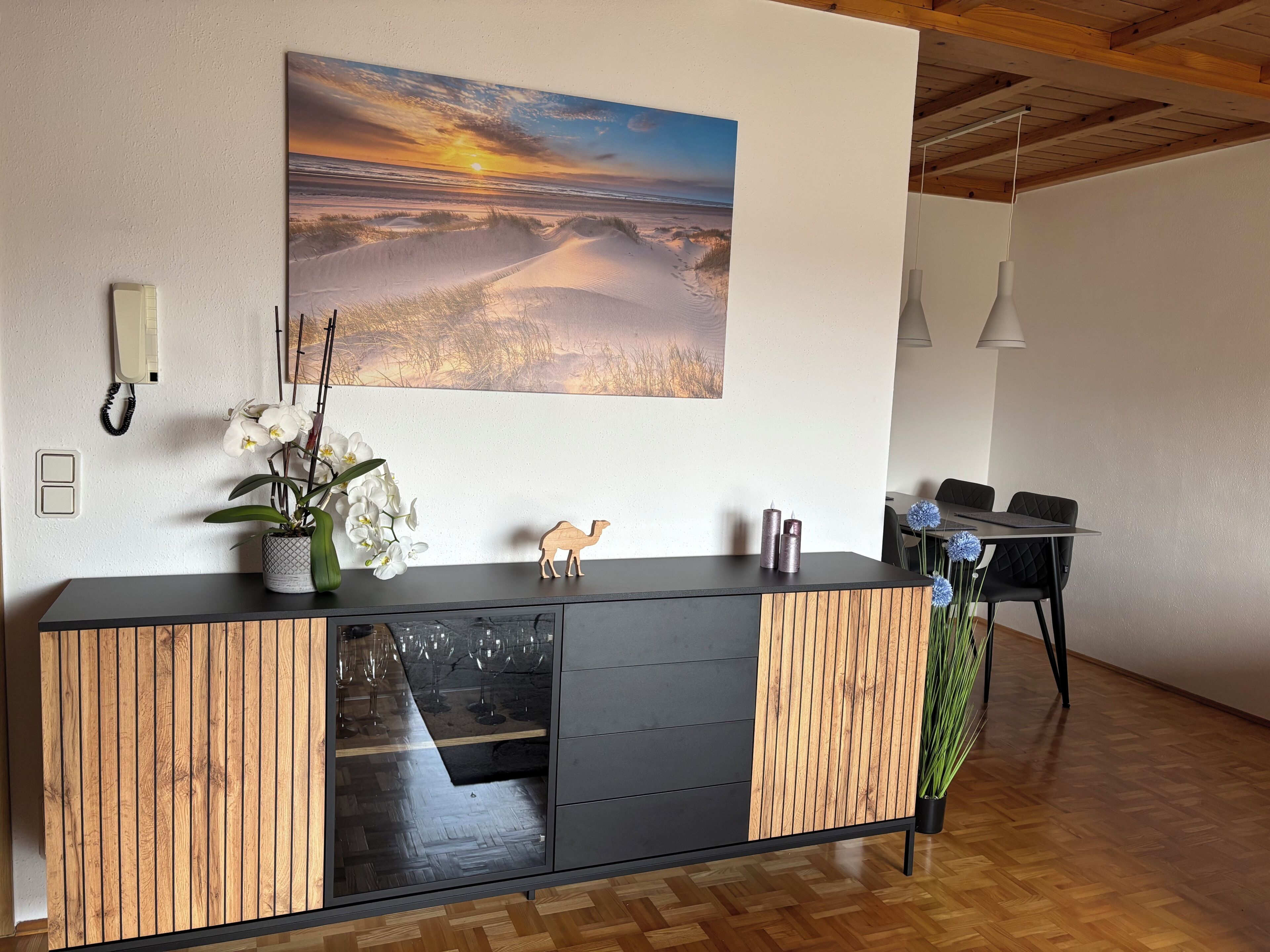 A modern reception area with a wooden and black console, a large landscape painting, a potted orchid, and a dining area with chairs and a table.