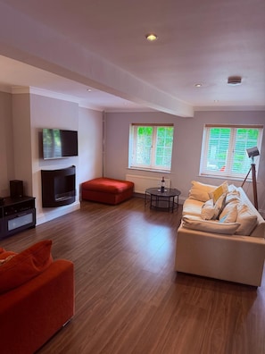 A modern living room with a flat-screen TV, a red ottoman, a beige sofa, and a small round table.