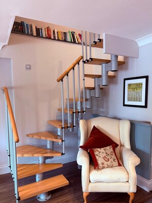 A modern staircase with a wooden finish and metal railings, a white armchair with a red cushion, and a bookshelf filled with books.