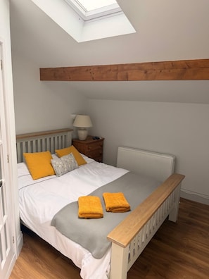 A bedroom with a skylight, wooden ceiling beams, a white radiator, a bedside table with a lamp, and two towels on the bed.