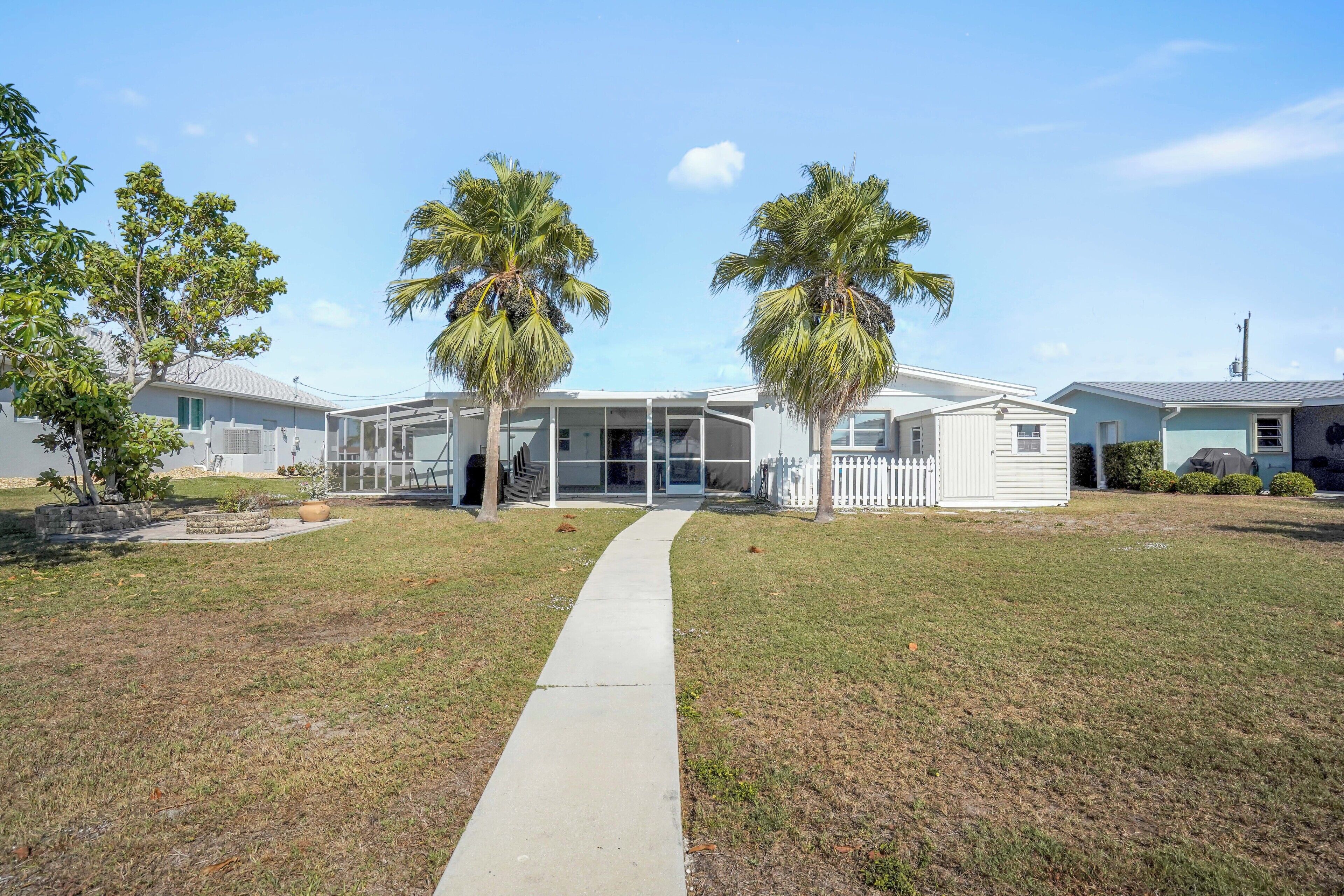 A residential area with a pathway leading to a house with a covered patio and two palm trees.