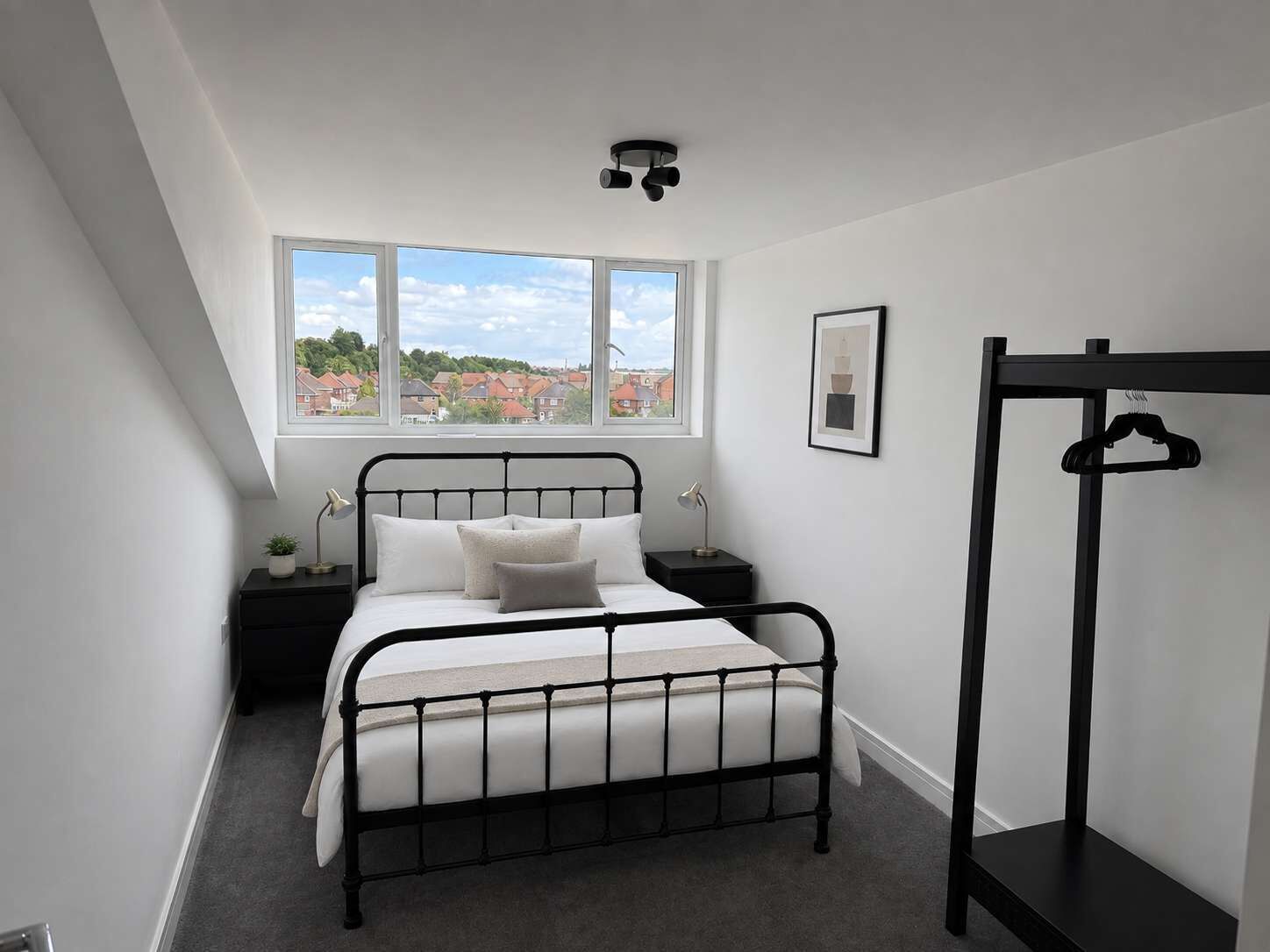 A bedroom with a bed, a window, a bedside table, a lamp, a picture frame, and a coat rack.
