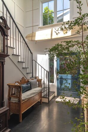 Un escalier avec un banc en bois à sa base, une porte bleue ornée d’une plante, et une vue sur la verdure à l’extérieur.