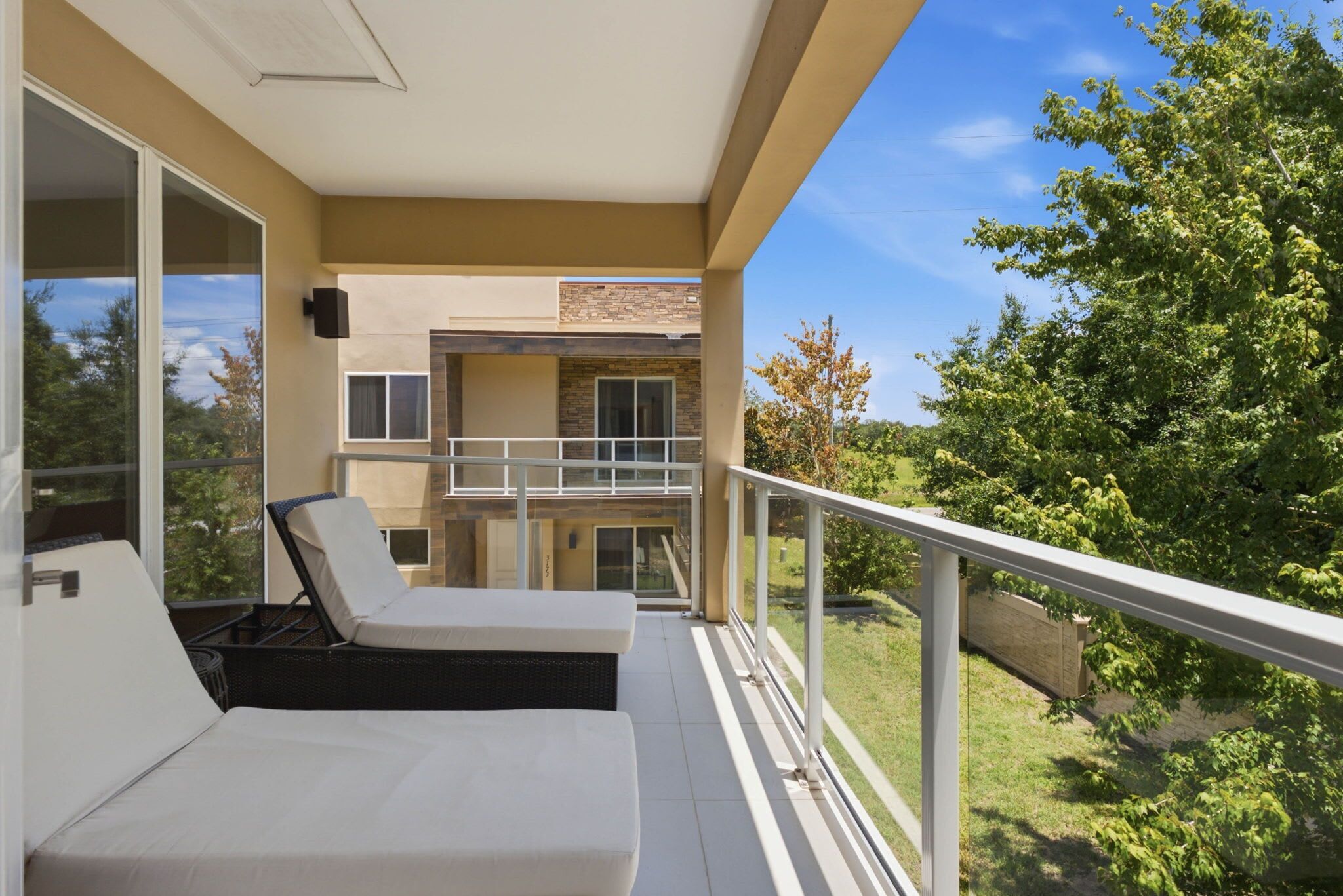 Uma varanda com área de estar acolchoada em branco, guarda-corpo de vidro e vista para uma paisagem verdejante.