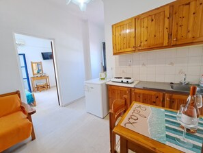 A compact kitchen with wooden cabinets, a white refrigerator, and a dining area with a table set for two. - Marvelous 1-bedroom apartment with AC in Αγια Πελαγια (ΑΓΙΑ ΠΕΛΑΓΙΑ)