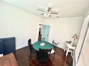 Uma sala de jantar com uma mesa redonda, quatro cadeiras, um ventilador de teto e uma porta que dá para outro cômodo.