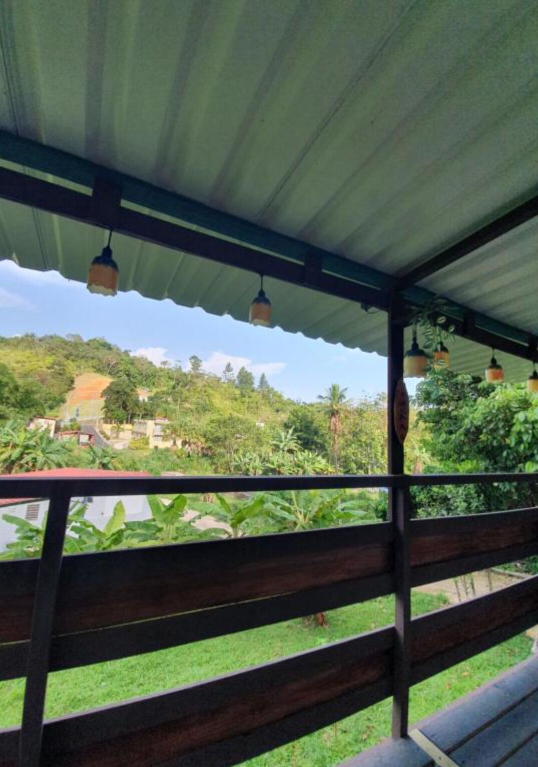 A covered outdoor area with a wooden bench and hanging lights, overlooking a lush green landscape with buildings in the distance.
