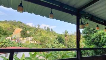 A covered outdoor area with a wooden bench and hanging lights, overlooking a lush green landscape with buildings in the distance.