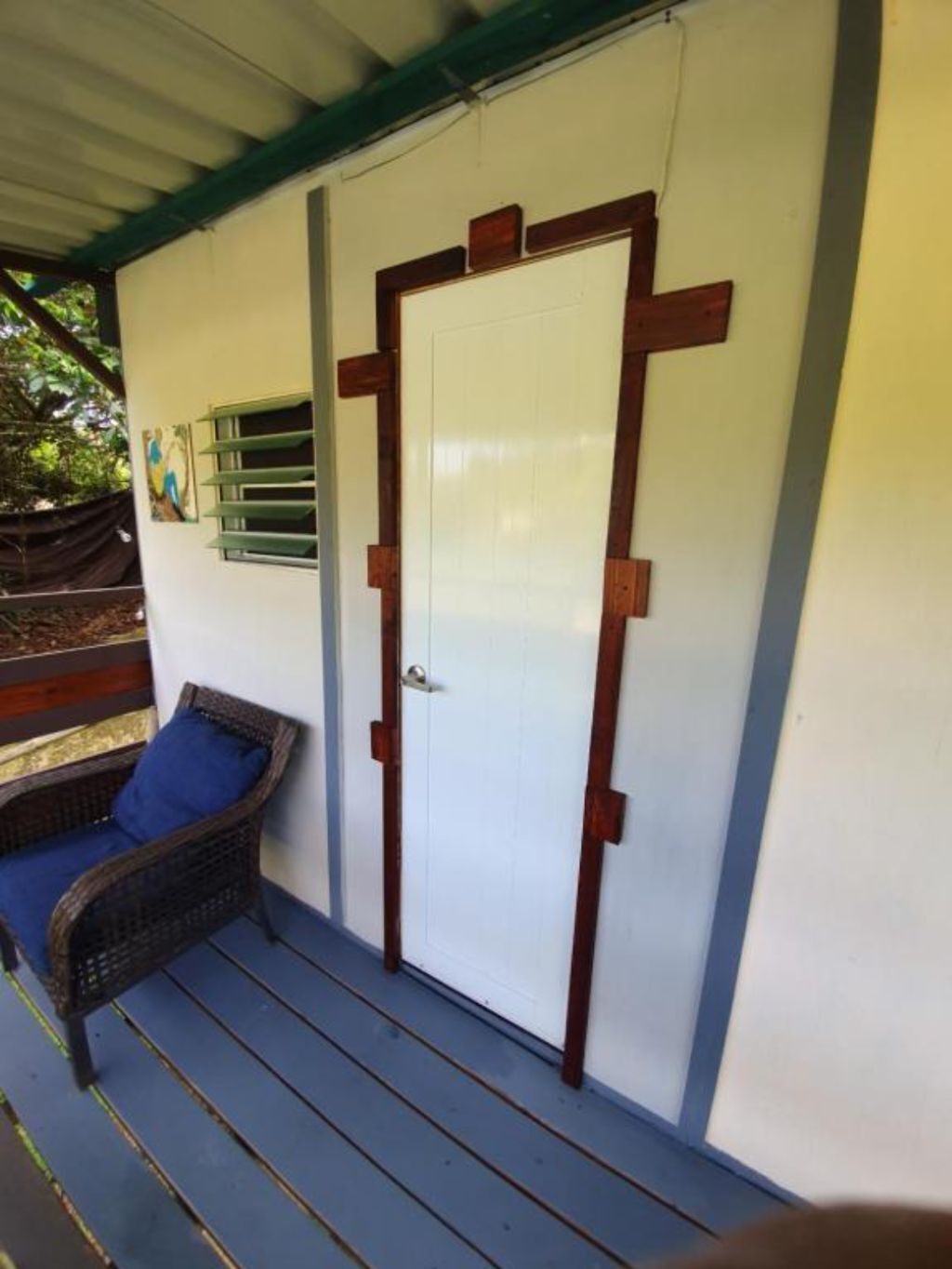 A covered porch with a white door, a wicker chair with a blue cushion, and a small window with horizontal blinds.