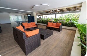 A modern lounge area with wicker furniture and orange cushions, a glass table, and a view of greenery through large windows. - Excentric at Juan Dolio Beach (Juan Dolio)