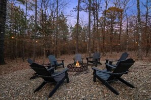 Ein Sitzbereich im Freien mit Adirondack-Stühlen, die um eine zentrale Feuerstelle angeordnet sind, umgeben von einer bewaldeten Landschaft.