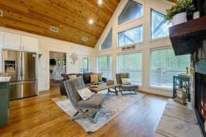 Un salón espacioso con techo de madera, ventanas amplias, chimenea de piedra y una cocina moderna con refrigerador de acero inoxidable.