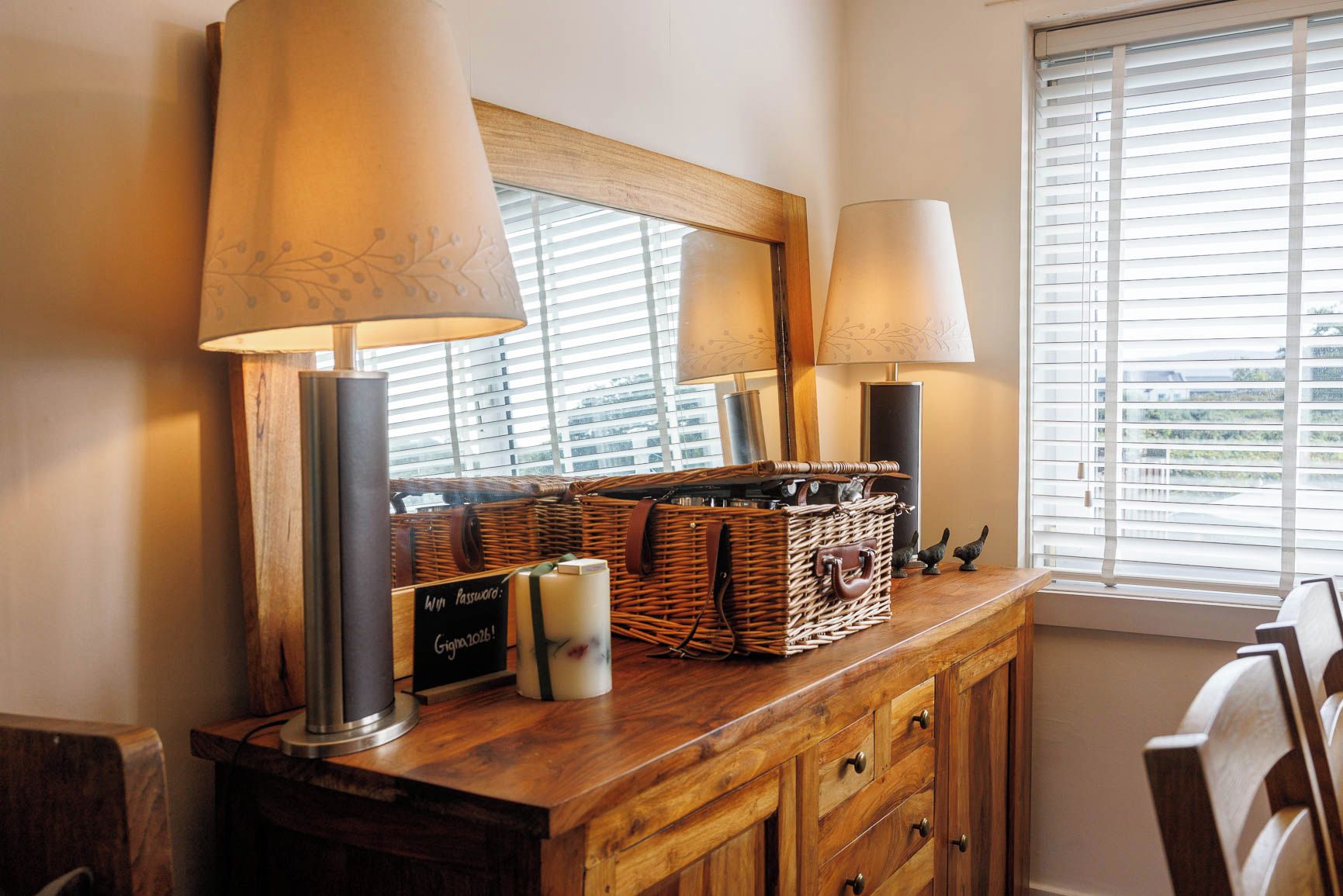 A wooden dresser with a mirror, two lamps, wicker baskets, and a sign.