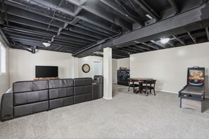 A basement with a black leather couch, a flat-screen TV, a clock, a table with chairs, and a pinball machine.