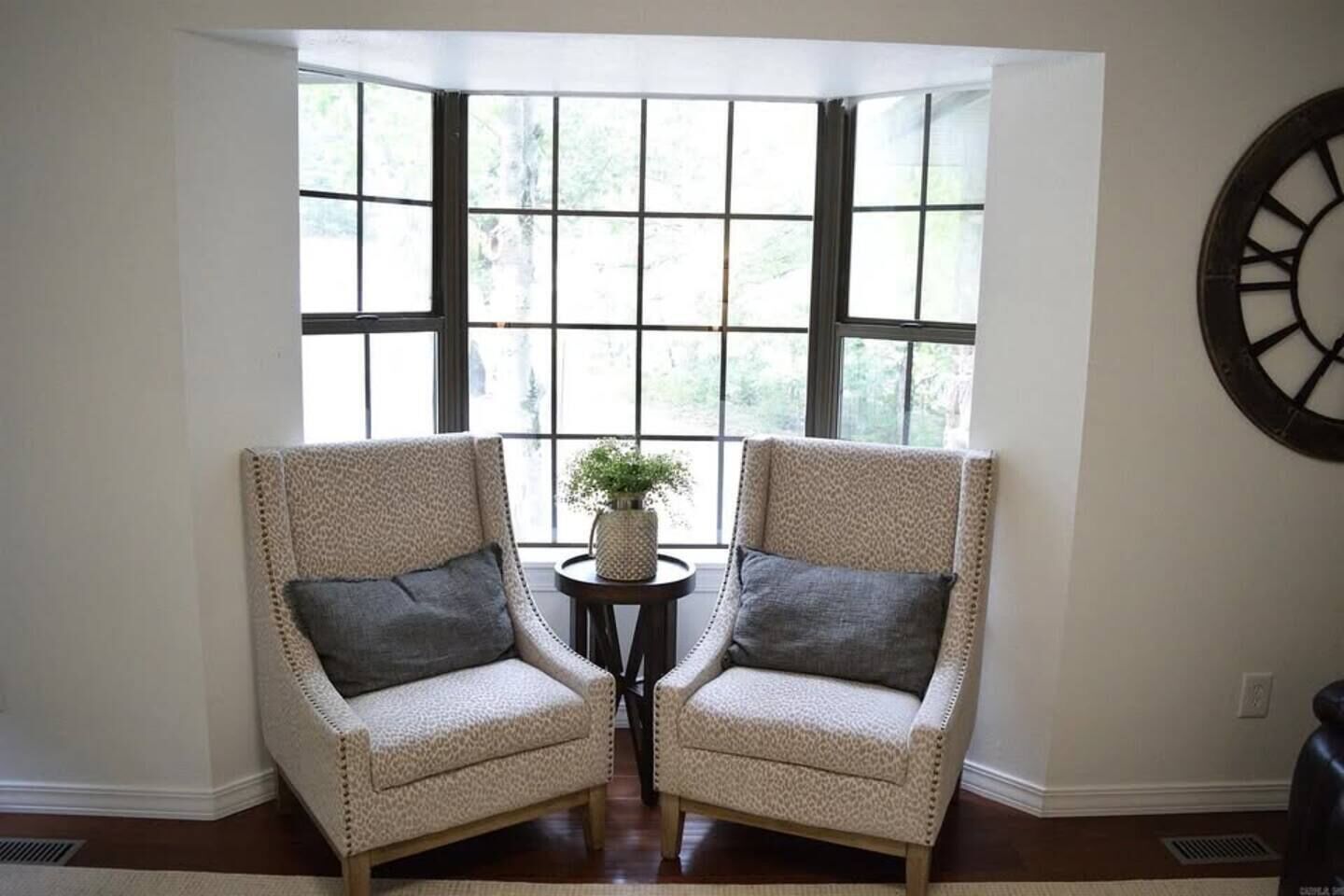 A corner room with two armchairs facing a window with multiple panes, a small round table with a plant, and a wall clock.