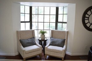 A corner room with two armchairs facing a window with multiple panes, a small round table with a plant, and a wall clock.