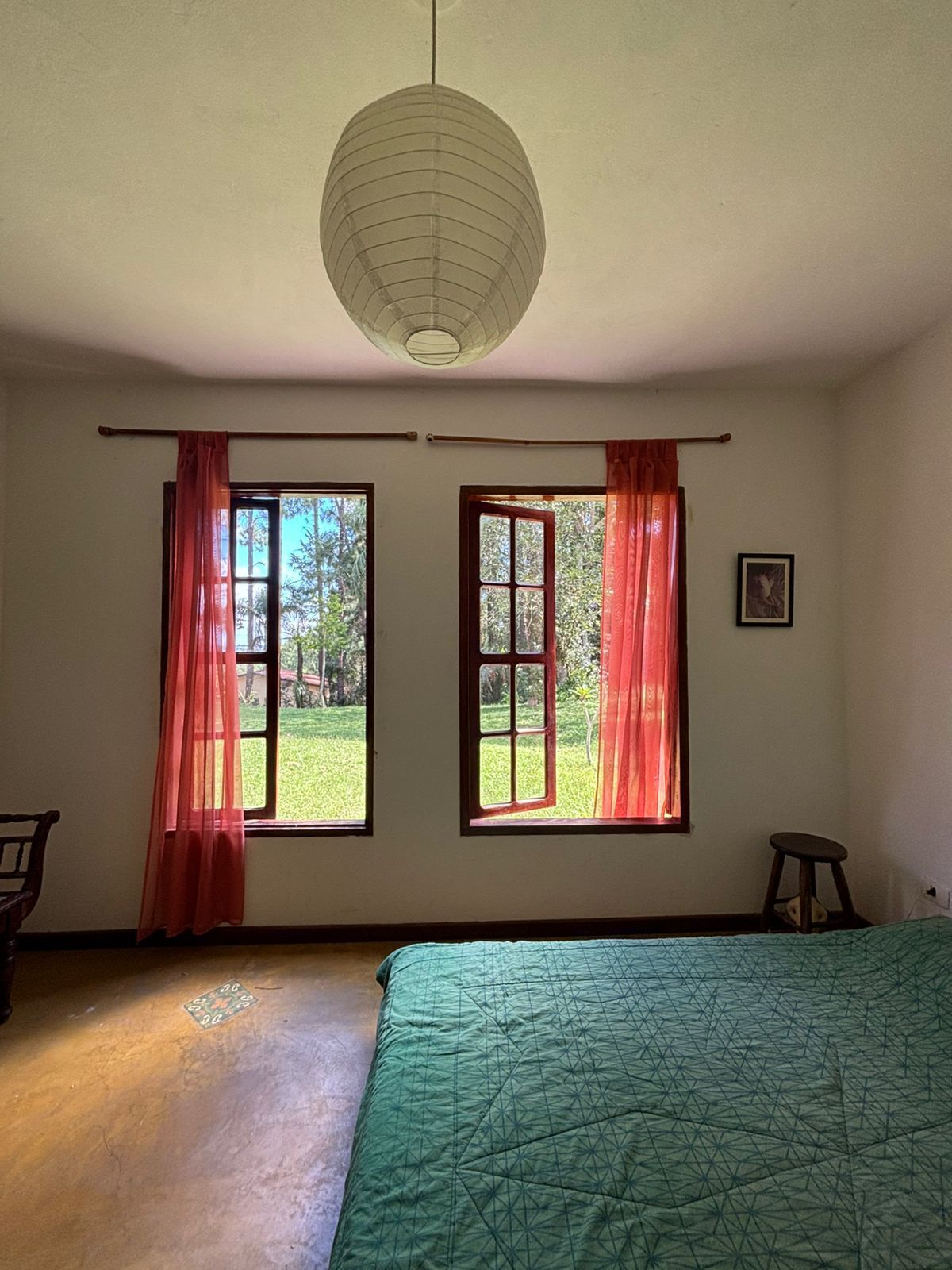 A room with a bed, two windows with curtains, a hanging light fixture, and a framed picture on the wall.