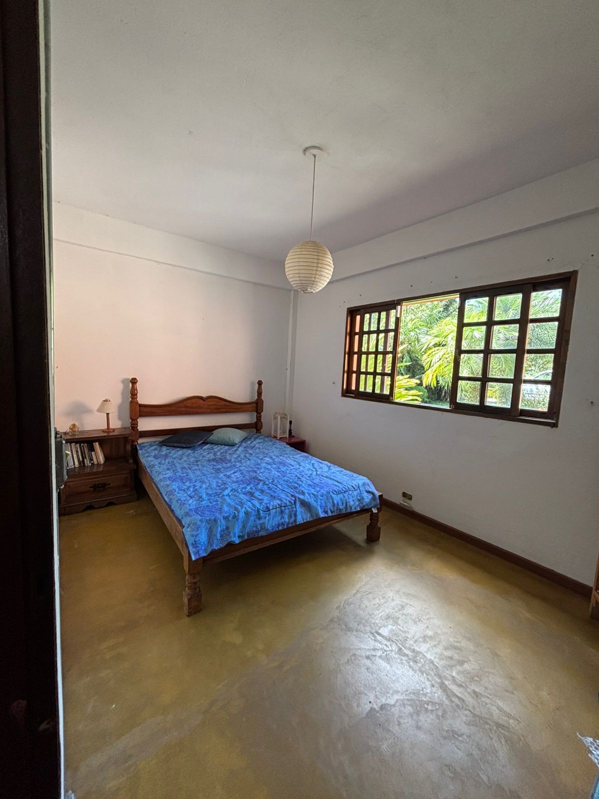 Un dormitorio con una cama individual, una mesita de noche, una ventana con vistas a zonas verdes y una lámpara colgante.