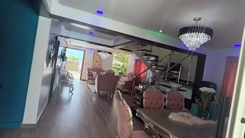 Une pièce spacieuse comprenant un coin repas avec une grande table et des chaises décorées, un lustre et un escalier menant à un niveau supérieur.