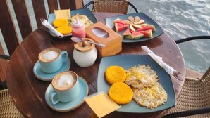 A table with breakfast items, including coffee, a wooden box, and plates with food, overlooking a beach with palm trees and the ocean.