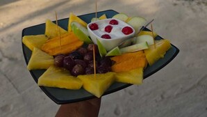 A plate of fruit and yogurt on a beach with palm trees and boats in the background.