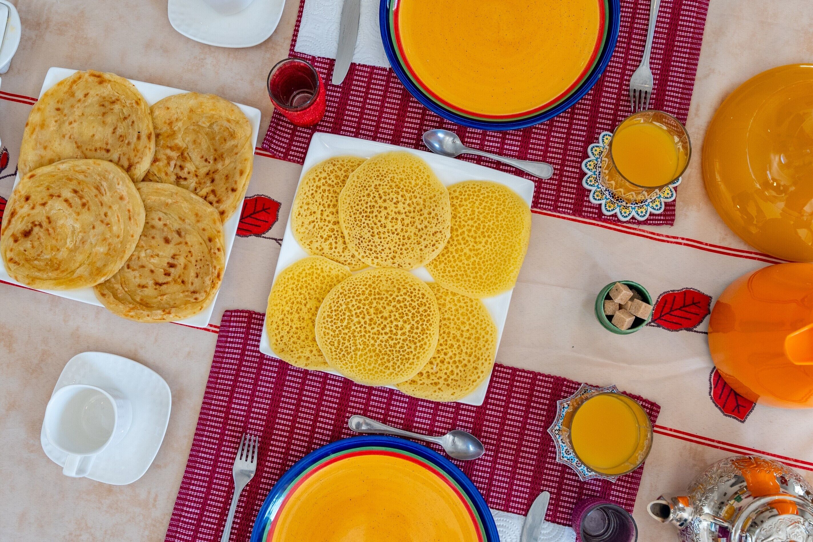 Een tafel gedekt met borden vol platbroden, een bord pannenkoeken en een karaf sinaasappelsap.