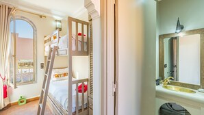 A bunk bed room with a ladder, a window with curtains, a bathroom with a sink and mirror, and a decorative wall piece.