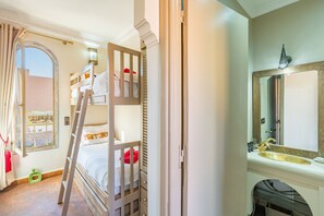 A bunk bed room with a ladder, a window with curtains, a bathroom with a sink and mirror, and a decorative wall piece.