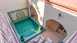 A rooftop pool with a jacuzzi, white outdoor furniture, and a decorative wall.