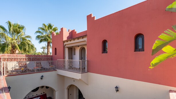 A building with a red facade, a balcony with ornate metal railings, and palm trees in the background.