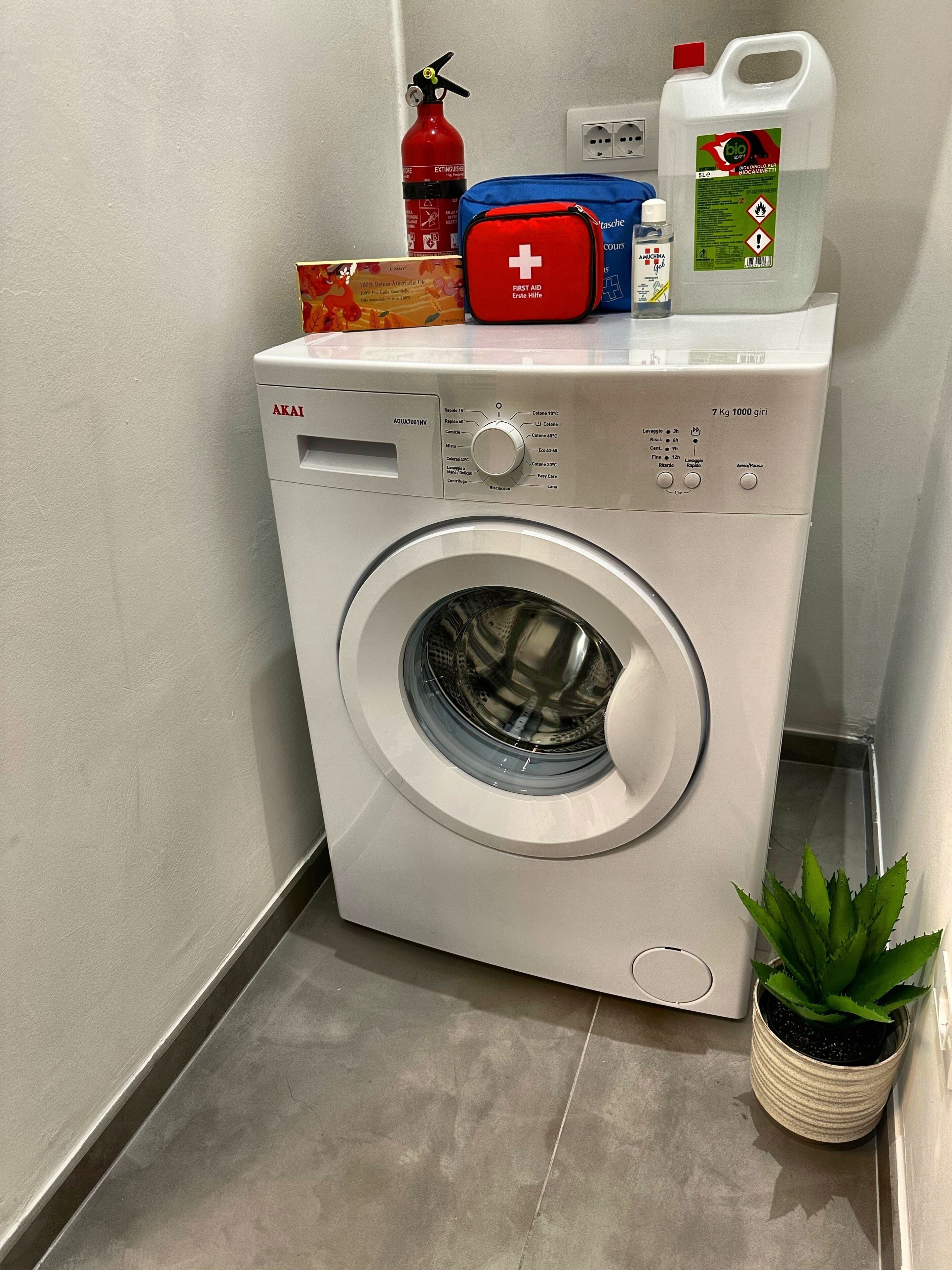 Una lavatrice a carica frontale con pannello di controllo, un estintore, un kit di pronto soccorso e una bottiglia di detergente.