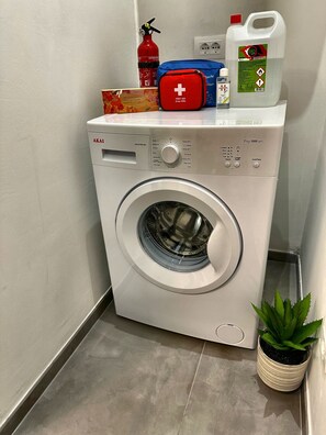 Una lavadora de carga frontal con panel de control, un extintor, un botiquín de primeros auxilios y una botella de solución limpiadora.