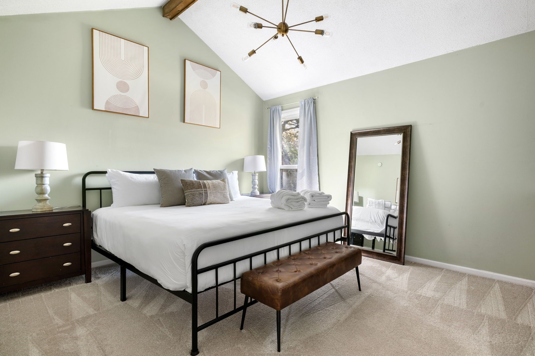 A bedroom with a bed, bedside tables, a bench, a mirror, and artwork on the walls.