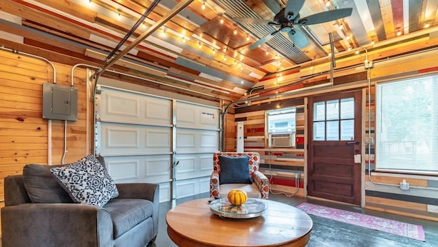 A room with a grey sofa, a round wooden table with a pumpkin, a ceiling fan, and a garage door.