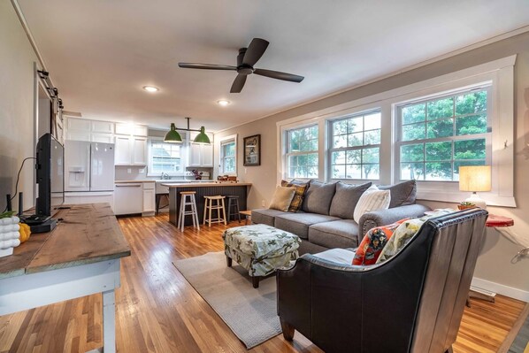 A modern kitchen with white cabinets, a dining area with stools, a grey sofa, and a black armchair.