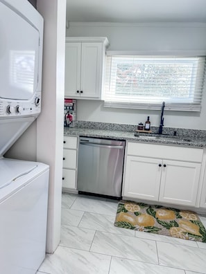 A kitchen with white cabinets, a stainless steel dishwasher, a granite countertop, and a window with blinds. - The Hampstead Suite- Free Parking & Walk to King! (Charleston)
