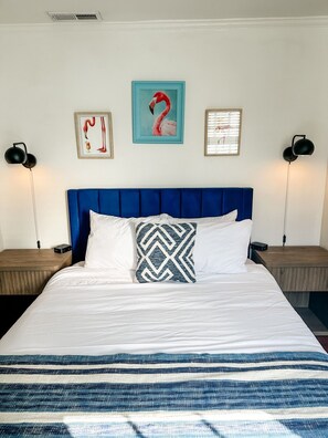 Ein Bett mit blauer Kopfteil, weißer Bettwäsche und einem gemusterten Kopfkissen. Es gibt zwei Nachttische mit Lampen, und ein Kunstwerk an der Wand über dem Bett.