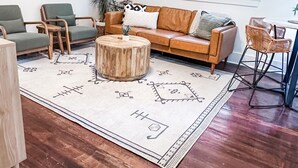 Ein Wohnzimmer mit Couch, zwei Sesseln, einem Holz-Couchtisch, einem Teppich mit Muster und einem großen Fenster mit Jalousien.
