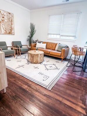 Ein Wohnzimmer mit Couch, zwei Sesseln, einem Holz-Couchtisch, einem Teppich mit Muster und einem großen Fenster mit Jalousien.