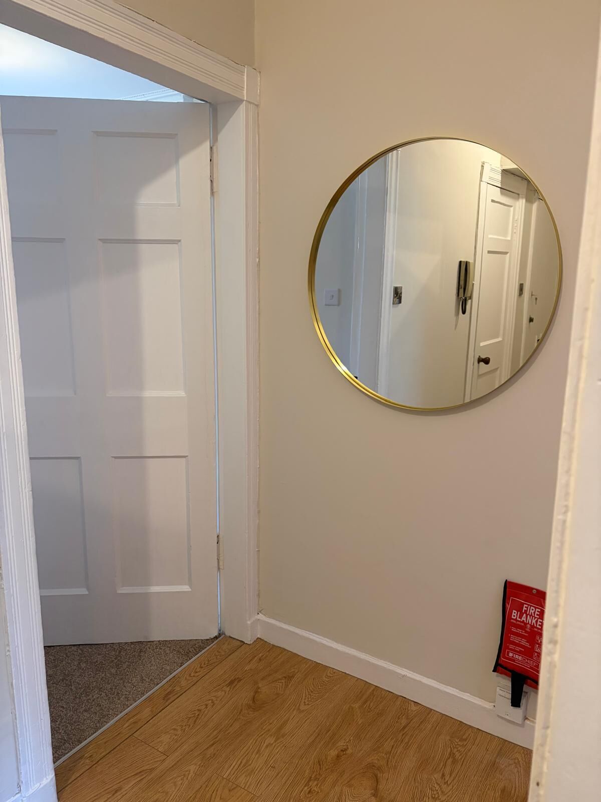 A hallway with a white door, a round mirror on the wall, and a fire blanket on the floor.