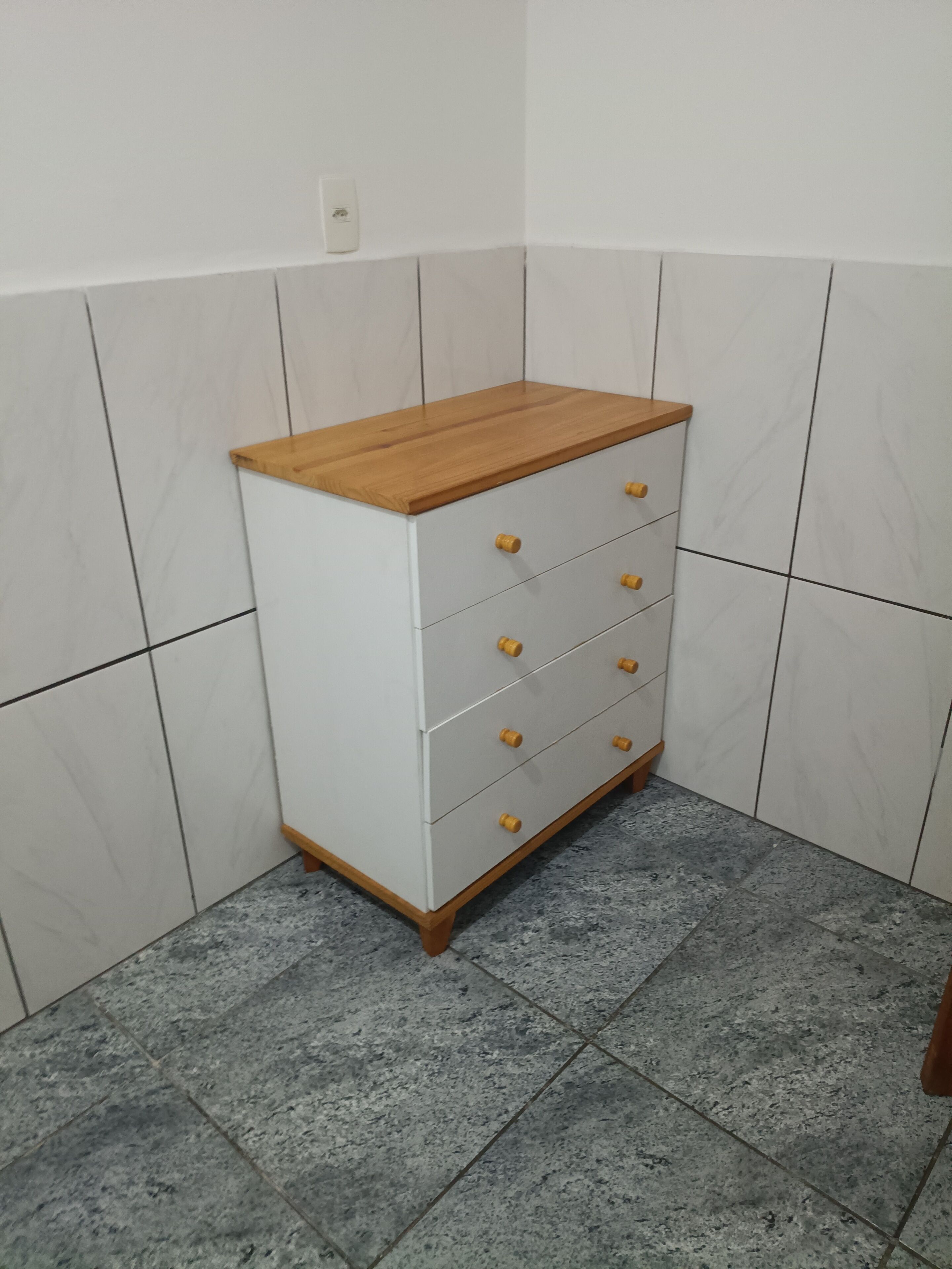 A white chest of drawers with wooden knobs and a wooden top, placed against a tiled wall in a corner.
