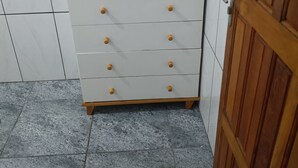 Uma cômoda branca com puxadores e tampo de madeira, posicionada em frente a uma parede revestida de azulejos com uma porta à direita.