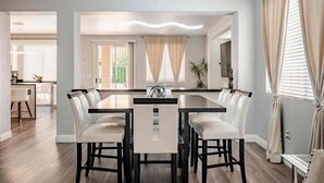A dining area with a long table and multiple chairs, a kitchen in the background, and a view of the outdoors through large windows. - 5BR Pool House + Billiards · Up to 15 Guests (Las Vegas)
